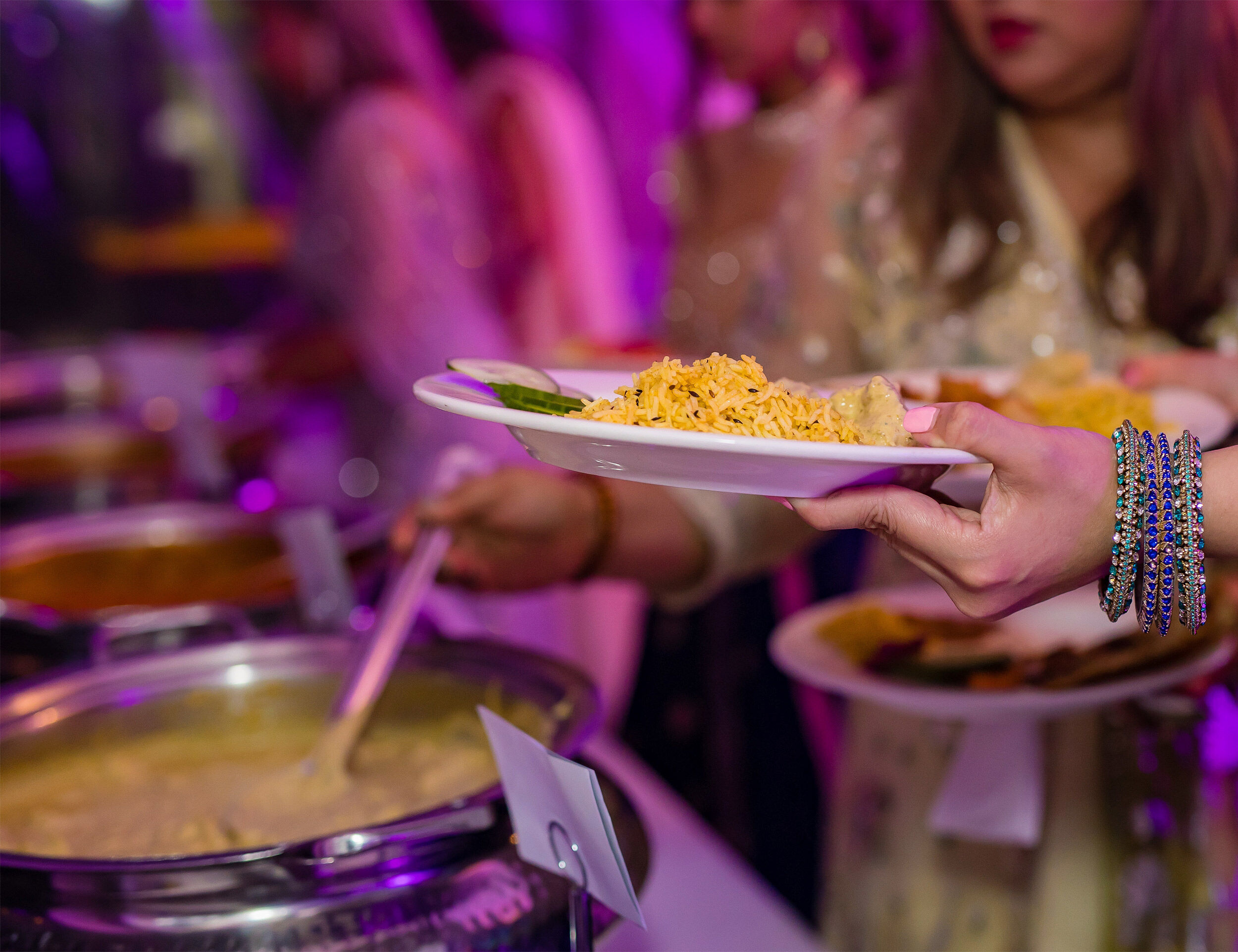 Guests serving rice and curry at a vibrant Indian wedding buffet under purple ambient lighting.